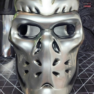 May include: A silver-colored hockey mask with a skull design and black eye holes. The mask has a textured surface and is made of plastic.