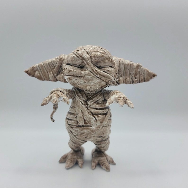 Puede incluir: Una peque&ntilde;a figura beige con forma de momia con un acabado texturizado y desgastado. La figura tiene orejas grandes, una boca ancha y est&aacute; envuelta en vendas.
