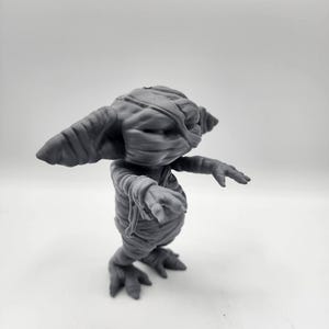 Puede incluir: Una figurita gris impresa en 3D de una criatura con apariencia de momia. La criatura tiene orejas grandes, un hocico largo y est&aacute; envuelta en vendas. Tiene cuatro patas y est&aacute; de pie sobre una superficie blanca.