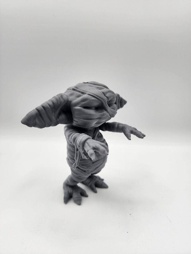 Puede incluir: Una figurita gris impresa en 3D de una criatura con apariencia de momia. La criatura tiene orejas grandes, un hocico largo y est&aacute; envuelta en vendas. Tiene cuatro patas y est&aacute; de pie sobre una superficie blanca.