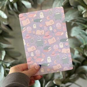 May include: A rectangular card with a lavender background featuring a repeating pattern of witch hats, books, potion bottles, ghosts, and crystal balls. The card is held in a hand, and the design is in pastel colors.