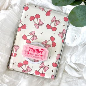 May include: Clear phone case with a pink cherry and bow pattern. A pink and white pop socket with the text "Thriller Book Club Lifetime Membership" is attached to the case. The case is on a white surface.