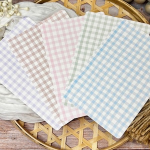 May include: A collection of square placemats in pastel colors, each with a classic gingham pattern. The placemats are arranged on a woven tray, with decorative elements like wooden beads and a tassel.
