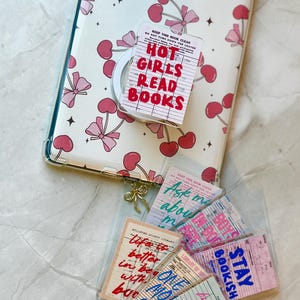 May include: A clear phone case with a cherry and bow pattern, a pop socket with "HOT GIRLS READ BOOKS" text, and several library card-themed stickers. The stickers have various phrases and designs.