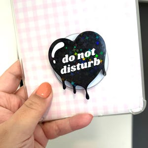 May include: A black heart-shaped phone grip with a holographic star pattern and white text that reads do not disturb. The heart design features a dripping liquid effect at the bottom edge and is attached to a clear phone case with a pink gingham insert.