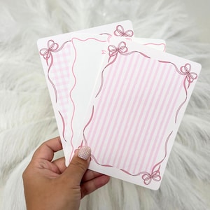 May include: Three white rectangular cards with pink ribbon and bow accents. One card has a pink gingham pattern, another has pink vertical stripes, and the third is plain. The cards are held against a white fluffy background.
