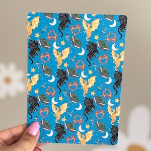 May include: A rectangular card with a blue background featuring a repeating pattern of black and gold dragons, books, hearts, moons, stars, and swords. The card is held by a hand with pink nail polish.