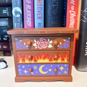 May include: A small brown three-drawer chest featuring decorative drawer fronts. The top drawer has pink roses and purple flowers, the middle drawer displays a yellow and red flame pattern, and the bottom drawer shows a yellow moon and stars.