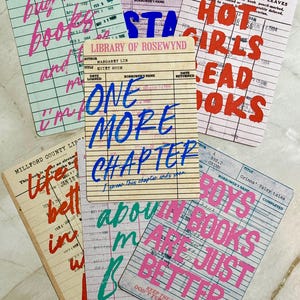 May include: A collection of vintage-style library cards with various handwritten phrases in pink, red, blue, and green. The cards feature text like "Buy me books," "Hot girls read books," and "Boys in books are just better."