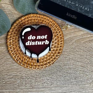 May include: A heart-shaped coaster with the words "do not disturb" in white lettering, featuring a dripping dark red design. The coaster sits on a woven rattan base, next to a Kindle e-reader.