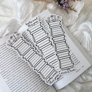 May include: White and black reading tracker bookmarks with the text "books read this year" and a stack of books design. The bookmarks are decorated with floral elements and hearts. The bookmarks are on top of an open book.
