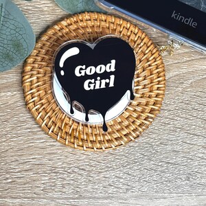 May include: A heart-shaped acrylic coaster with the words "Good Girl" in white on a black background. The coaster has a dripping design and sits on a woven rattan coaster. A Kindle e-reader is visible in the background.