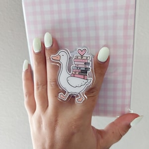 May include: A clear acrylic sticker featuring a white goose carrying a stack of books with pink spines and a heart. The goose has an orange beak and feet. The background is a pink and white gingham pattern.