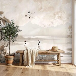 May include: A wooden bench with a beige knit throw blanket and a straw hat, positioned in front of a wall mural depicting a landscape with two cranes, mountains, and trees. A potted olive tree stands to the left of the bench on a wooden floor.
