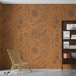Puede incluir: Una habitación con paredes cubiertas con un patrón floral repetitivo en tonos marrón y cobre. Una silla de alambre dorada está en la esquina, y una estantería de color marrón oscuro está contra la pared, con libros y objetos decorativos.