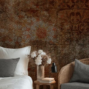 May include: A bedroom scene with a textured, rusty brown wall. A bed with white linens and a checkered pillow is on the left. A wicker chair with a matching pillow is on the right. A small wooden table holds a vase of white flowers and a black lamp.