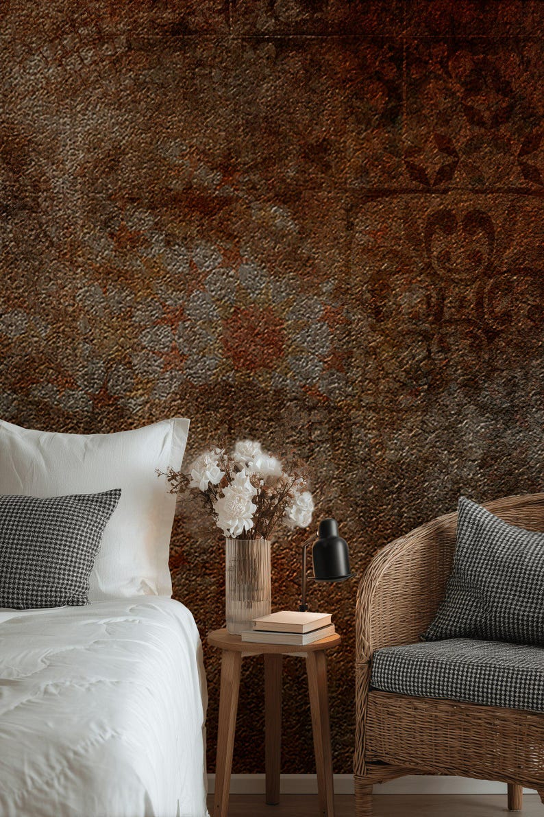 May include: A bedroom scene with a textured, rusty brown wall. A bed with white linens and a checkered pillow is on the left. A wicker chair with a matching pillow is on the right. A small wooden table holds a vase of white flowers and a black lamp.