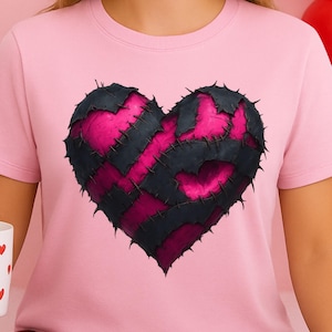 May include: A pink t-shirt featuring a large heart design. The heart is composed of black and pink patches, stitched together. The person is also holding a white mug with red heart designs. Red balloons and heart-shaped decorations are in the background.