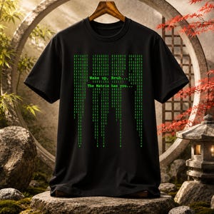 May include: A black short-sleeve t-shirt featuring a green digital code pattern printed on the front. The graphic includes the text "Wake up, Bruh... The Matrix has you..." written in a digital font style, mimicking falling computer code.