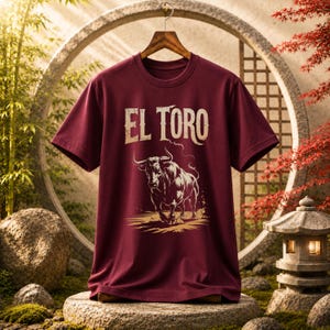 Peut inclure: Un t-shirt &agrave; manches courtes bordeaux avec une impression graphique repr&eacute;sentant un taureau en charge et le texte "EL TORO" dans une police claire et vieillie. Le t-shirt est pr&eacute;sent&eacute; sur un cintre en bois devant un cadre circulaire.