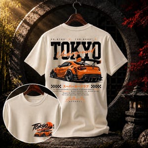 May include: A cream-colored t-shirt featuring a graphic of an orange sports car on the back with the text TOKYO Underground, coordinates, and Japanese characters. The front includes a smaller graphic of the same car and the text Tokyo Underground.