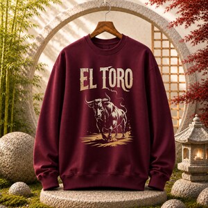 Peut inclure: Un sweat-shirt &agrave; col rond et manches longues de couleur bordeaux, orn&eacute; d'une impression graphique repr&eacute;sentant un taureau en position de charge. Au-dessus du taureau, le texte EL TORO est imprim&eacute; dans une police blanc cass&eacute; vieillie.