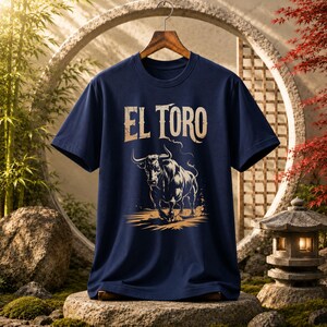 Peut inclure: Un t-shirt bleu marine &agrave; manches courtes avec une impression graphique d'un taureau en charge de couleur beige clair. Au-dessus du taureau, le texte EL TORO est imprim&eacute; dans une police beige clair vieillie. Le t-shirt est sur un cintre en bois.