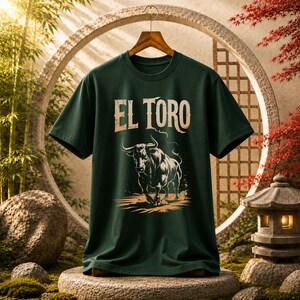 Peut inclure: Un t-shirt &agrave; manches courtes vert fonc&eacute; avec une impression graphique d'un taureau en charge en noir et blanc. Au-dessus du taureau, le texte EL TORO est imprim&eacute; dans une police de couleur claire et vieillie. Le t-shirt a un col rond.