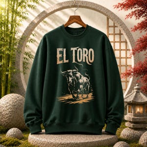 Peut inclure: Un sweat-shirt &agrave; col rond vert fonc&eacute; avec une impression graphique repr&eacute;sentant un taureau en charge dans une palette de couleurs beige et noir. Au-dessus du taureau, le texte EL TORO est imprim&eacute; dans une police serif grasse et vieillie.