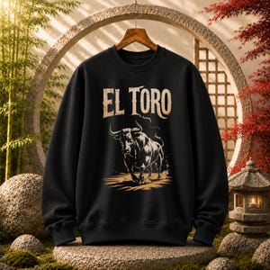 Peut inclure: Un sweat-shirt noir &agrave; manches longues avec une impression graphique repr&eacute;sentant un taureau en position de charge. Au-dessus du taureau, le texte EL TORO est imprim&eacute; dans une police beige clair vieillie, avec des d&eacute;tails de poussi&egrave;re.