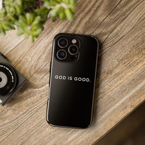 May include: A black phone case with the text "GOD IS GOOD" in white. The phone case is on a wooden surface, with a camera and a plant in the background.