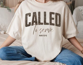 Può includere: T-shirt color crema con la scritta "CALLED to serve" in marrone scuro. Il testo "MARK 10:45" è sotto. La maglietta, dalla vestibilità ampia, è abbinata a jeans blu. L'insieme ha un aspetto rilassato.
