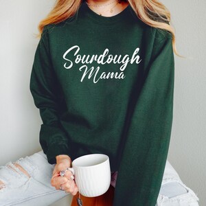 Może przedstawiać: Ciemnozielona bluza z napisem "Sourdough Mama" w białym skrypcie. Bluza ma okrągły dekolt. Osoba trzyma biały kubek.