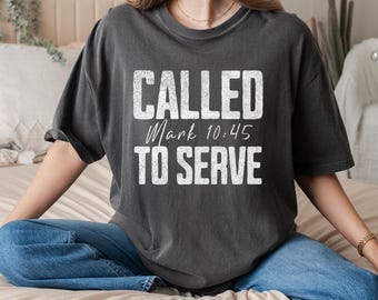 Può includere: T-shirt grigio scuro con la scritta "CALLED Mark 10:45 TO SERVE" in lettere bianche. La maglietta ha maniche corte e una vestibilità ampia. La persona indossa jeans blu.