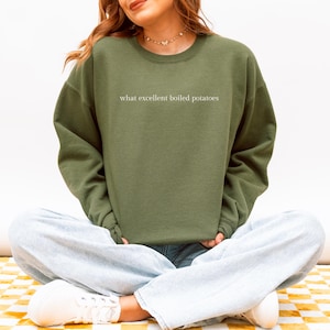Op de afbeelding: Olijfgroene sweatshirt met de witte tekst "what excellent boiled potatoes". De ronde hals wordt gedragen met een lichtblauwe jeans en witte sneakers. De persoon zit met gekruiste benen.