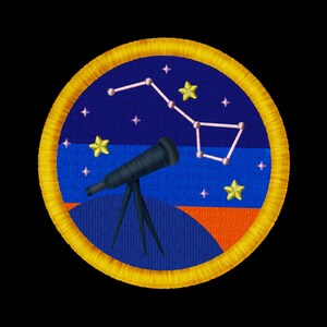 May include: A circular embroidered patch featuring a dark blue night sky with a light pink constellation, yellow stars, and a black telescope on a tripod. The background includes a blue horizon line and an orange base, all bordered by a thick yellow thread.