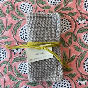 May include: A small, hand-knitted gray wool dishcloth with a yellow string tied around it. The dishcloth is on a pink and green floral fabric background. A white tag is attached to the string with the text "100% Wool LeftyHandmade.Etsy.com".