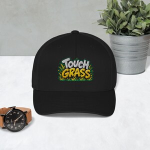 Puede incluir: Una gorra de béisbol negra con un gráfico bordado en el panel frontal. El diseño muestra el texto "TOUCH GRASS" en letras blancas y amarillas, rodeado de briznas de hierba verdes estilizadas. La gorra tiene una corona estructurada y visera.