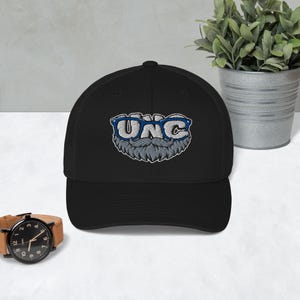 Puede incluir: Una gorra de béisbol negra con un bordado de las letras UNC en blanco, acentuado por un par de gafas azules y un bigote gris. El diseño está centrado en el panel frontal de la gorra, que cuenta con una corona estructurada y una visera curva.