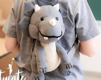 May include: A gray dinosaur-shaped backpack with white horns and a white nose is worn by a person. The backpack has a smiling face and a beige belly. The straps are gray. The word "Artucate" is written in white on the bottom left.