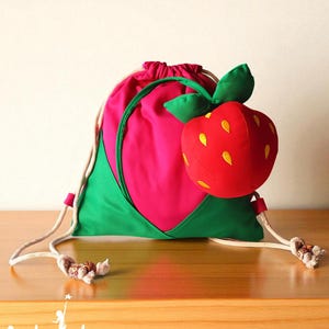 Puede incluir: Una mochila con cordón en diseño de fresa. La bolsa es principalmente rosa con detalles verdes y una gran fresa de peluche adjunta. La fresa tiene detalles de semillas amarillas y hojas verdes. La bolsa tiene cordones blancos. La palabra "Artucate" está en la parte inferior.