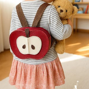 Op de afbeelding: Een rode appelvormige rugzak met een crèmekleurig appelplakontwerp en bruine bandjes. De rugzak wordt gedragen door een kind dat een gestreept shirt en een roze rok draagt. Een teddybeer wordt in de armen van het kind gehouden. De merknaam "Artucate" is zichtbaar.