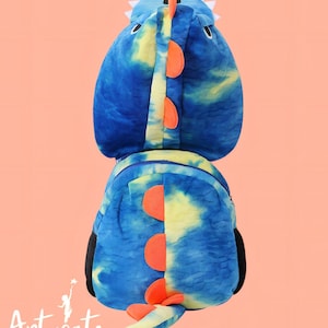 Puede incluir: Una mochila de dinosaurio azul y amarilla teñida con detalles en naranja. La mochila tiene una capucha con forma de cabeza de dinosaurio con púas blancas y una cola. Se ven paneles laterales negros y la marca "Artucate".