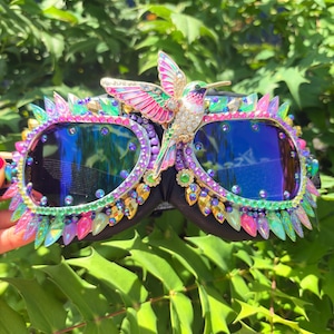 May include: Goggles featuring dark blue reflective lenses, heavily embellished with a vibrant array of multi-coloured crystals and iridescent spikes. A prominent, detailed hummingbird ornament with pink, green, and white accents sits centred.