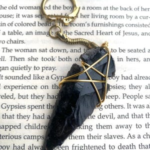 May include: A black arrowhead pendant, wrapped in gold-coloured wire with a five-pointed star design. The pendant is attached to a gold-coloured keyring. The background is a book with printed text.