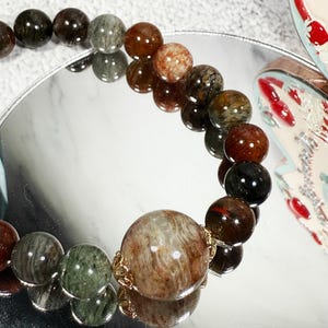 May include: A beaded bracelet featuring a variety of earth-toned, round gemstones. The beads range in color from deep brown and black to green and reddish-brown. The bracelet is displayed on a reflective surface.