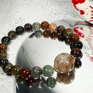 May include: A beaded bracelet featuring a variety of earth-toned gemstones. The bracelet includes a large, round bead with gold-colored accents. The beads are in shades of brown, green, and red, reflecting light.
