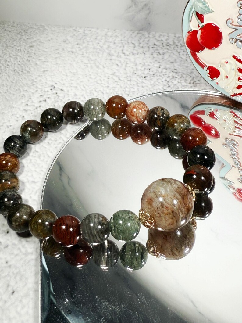 May include: A beaded bracelet featuring a variety of earth-toned gemstones. The bracelet includes a large, round bead with gold-colored accents. The beads are in shades of brown, green, and red, reflecting light.