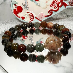 May include: A beaded bracelet featuring a mix of dark green, brown, and black round beads. The bracelet has a large, round, light brown bead with gold-colored accents. The bracelet is displayed on a reflective surface.
