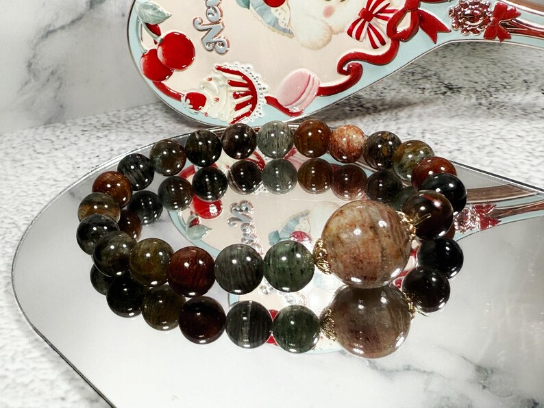 May include: A beaded bracelet featuring a mix of dark green, brown, and black round beads. The bracelet has a large, round, light brown bead with gold-colored accents. The bracelet is displayed on a reflective surface.